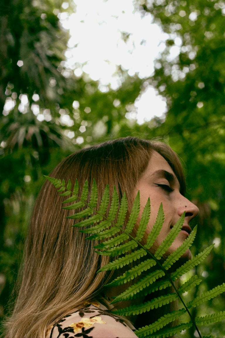Woman in profile with long brown hair framed by lush green fern fronds in a forest setting with dappled sunlight