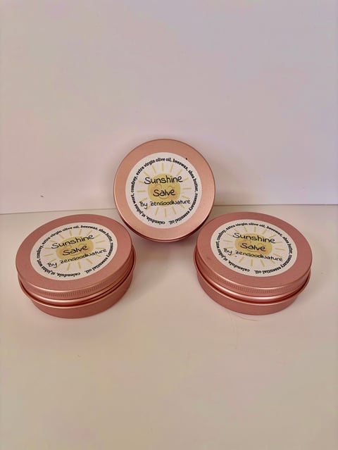Three rose-colored metal tins of Sunshine Salve with cream-colored labels arranged on a white surface