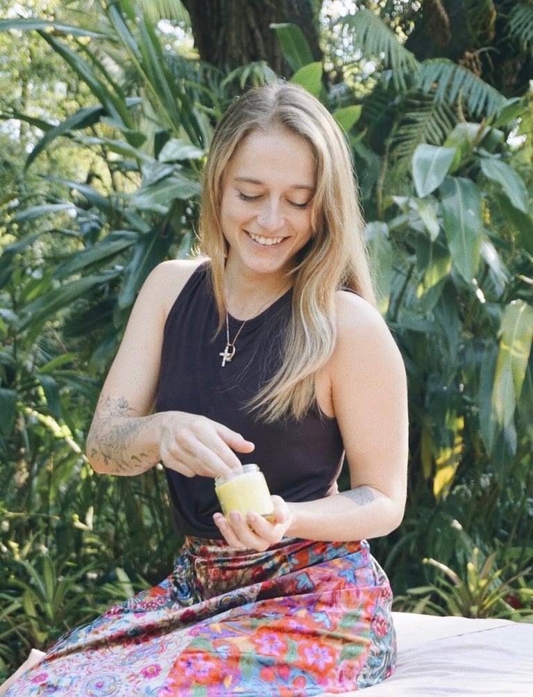 Woman with blonde hair in black tank top and colorful pants smiling while holding fruit near tropical plants