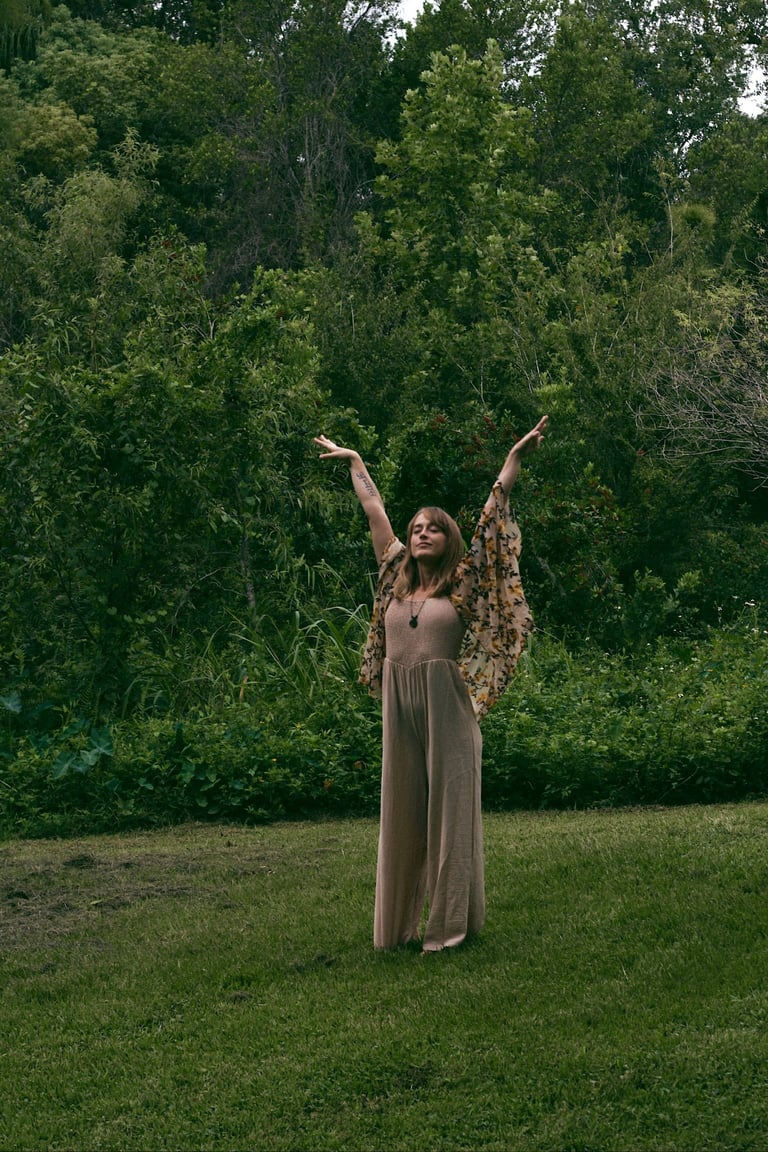 Woman in beige dress with arms raised standing on grass in front of lush green trees